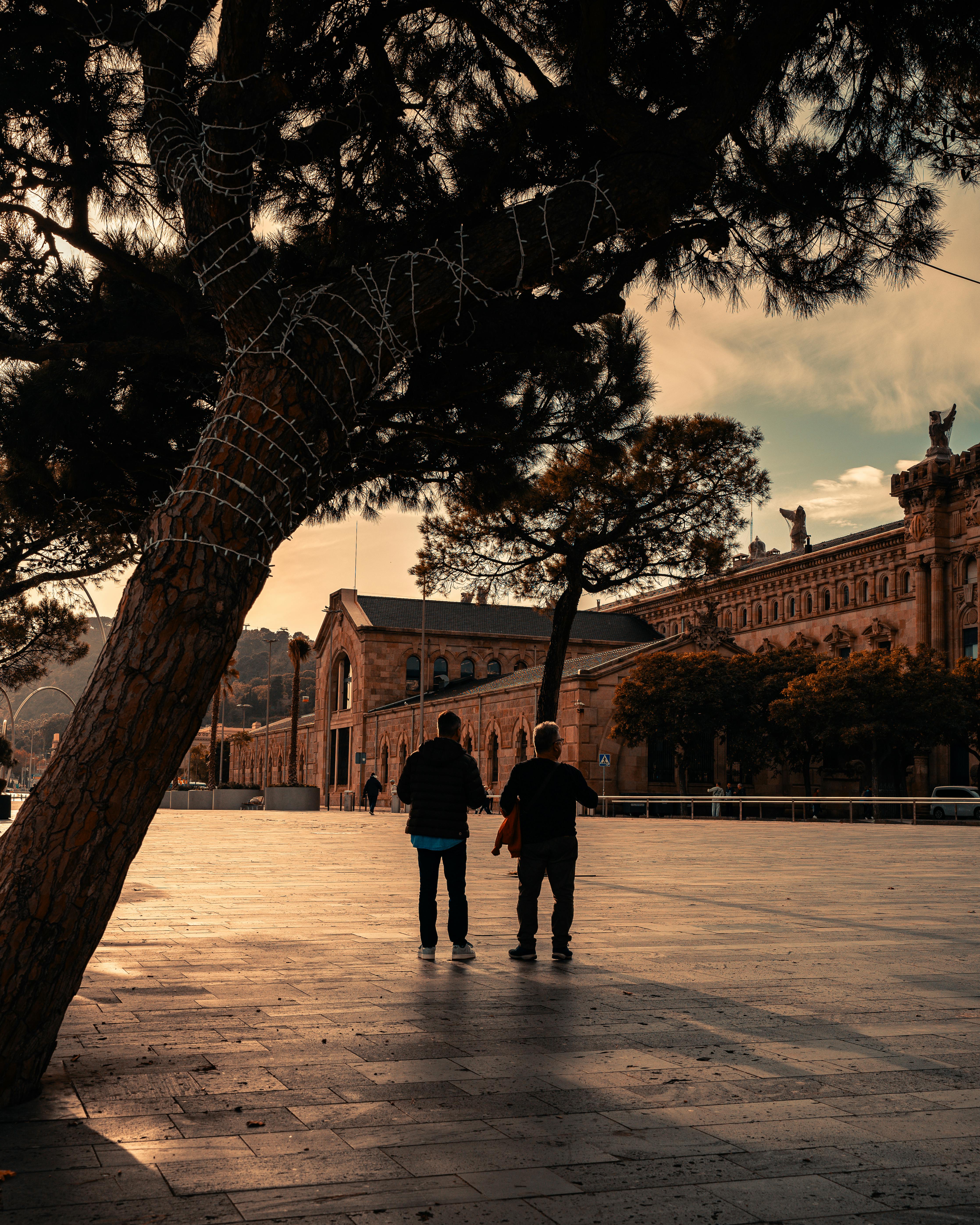 Free Two individuals stroll in a sunlit plaza near a historic building in Barcelona. Stock Photo